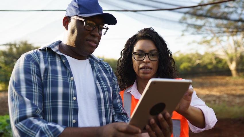 A farmer stands outside holding a tablet as a woman assists him with using it.