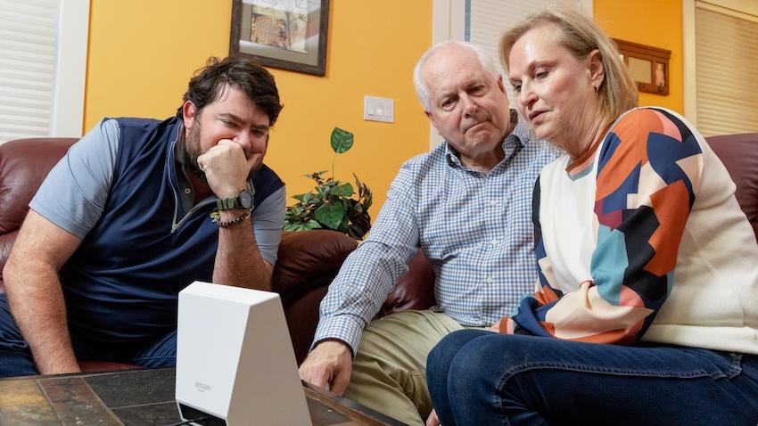 An older couple sit on a sofa as a younger man guides them through using Amazon Alexa